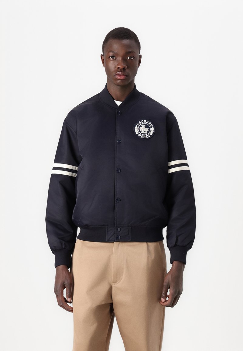 Chaqueta bomber en color navy con un logo en el pecho, detalles en blanco en los puños y cierre de botones, fabricada con un tejido liso y duradero.