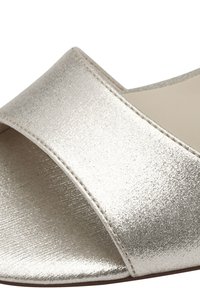 Sandale à glissière en argent métallique avec une large sangle croisée, texture lisse et design minimaliste. L'intérieur est d'une couleur neutre contrastante.
