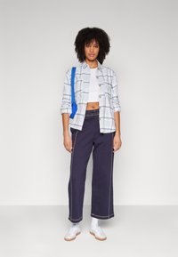 Lichtblauw geruit overhemd over een wit crop top, gecombineerd met donkerblauwe wijde broek met contrasterende stiksels en witte sneakers.