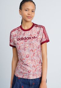 Adidas rozā un sarkanā ziedu raksta T-krekls ar apaļu kakla izgriezumu, tumši sarkaniem akcentiem un trim svītrām uz piedurknēm. Izgatavots no viegla auduma.