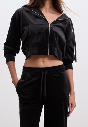 Frau trägt eine schwarze, kurze Zip-up-Hoodie und passende schwarze Hosen mit Kordelzug, steht mit einer Hand in der Tasche vor einfarbigem Hintergrund.