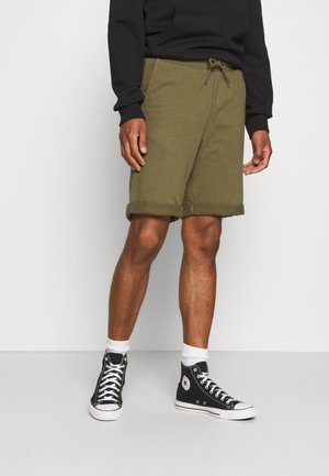 Mann trägt olivgrüne Shorts mit Kordelzug, schwarze High-Top-Sneakers mit weißen Sohlen und Socken, steht vor weißem Hintergrund.