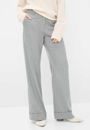 Femme portant un pantalon large gris clair à fines rayures avec revers aux ourlets, associé à un pull crème et des chaussures blanches pointues.