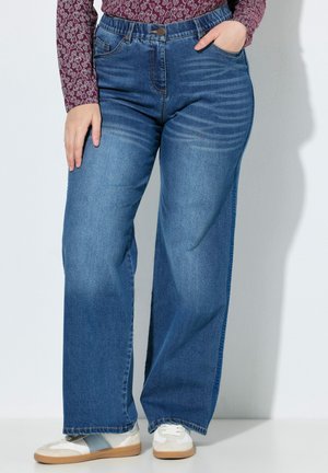 Blå jeans med vide ben båret med hvide og grå sneakers, kombineret med en langærmet mønstret top, hænderne hvilende på hofter og lår.