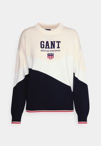 Pull en maille crème, marine et blanc arborant un logo "GANT" audacieux, un emblème de sportwear et des poignets côtelés avec des accents rouges. Coupe classique.