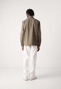 Brown lightweight jacket with a relaxed fit, buttoned cuffs, and white loose-fitting trousers, paired with light-colored sneakers.