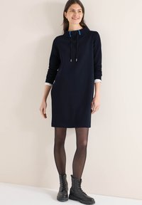 Robe sweat-shirt bleu marine avec un col montant, des cordons de serrage bleus contrastants et des manches longues. Portée avec des bottes noires à lacets et des collants transparents.