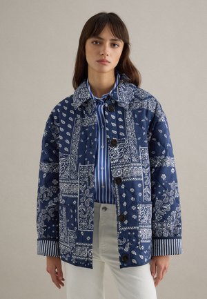 Junge Frau trägt eine blau gemusterte gesteppte Jacke über einem blau-weiß gestreiften Hemd, kombiniert mit weißen Hosen, stehend vor einem einfarbigen Hintergrund.