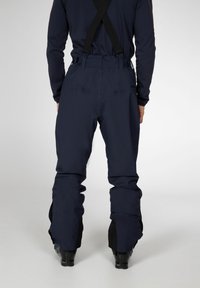 Skibroek in donker marineblauwe stof met verstelbare bretels, losse pasvorm, versterkte boorden en opvallende naaddetails. Zwarte accenten aan de onderkant.