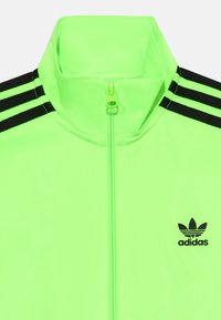 Neon zeleni zip-up jakna s visokim ovratnikom, koja ima tri crne pruge na svakom ramenu i mali crni Adidas logo na prsima.