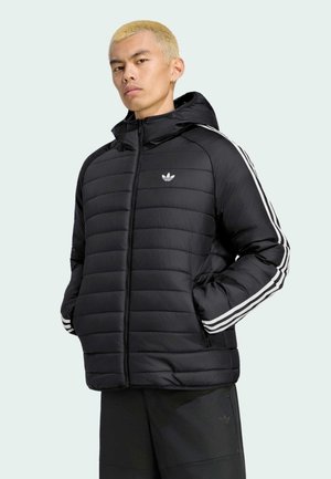 Mand med kort blondt hår iført sort polstret Adidas jakke med hvide striber på ærmerne, stående med hænderne i lommerne.