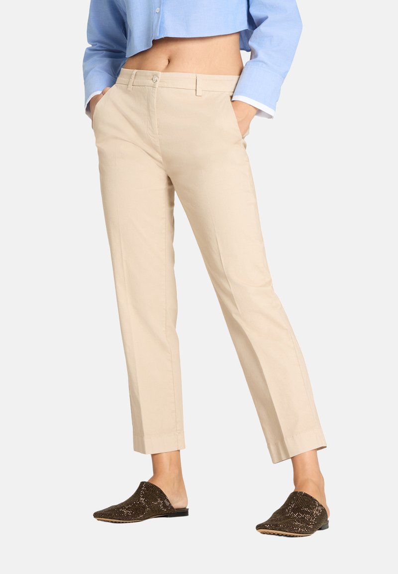 Donna con pantaloni beige a gamba dritta, camicia azzurro chiaro corta e scarpe slip-on marrone scuro con decorazioni, in piedi con le mani nelle tasche.