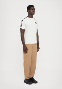 White t-shirt with black accent stripes on sleeves, paired with tan cargo pants featuring side pockets. Black shoes complete the look.