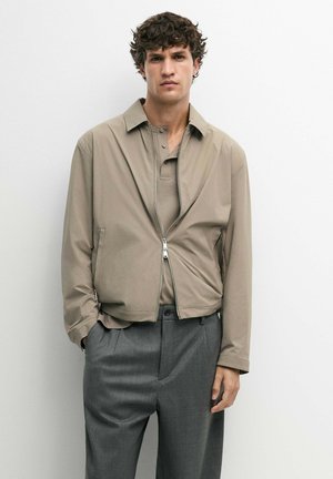 Mand med krøllet mørkt hår iført en beige lynlåstrøje over en beige skjorte med krave og grå bukser, stående mod en hvid væg.