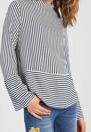 Vrouw draagt een navyblauw-wit gestreept shirt met wijde mouwen en blauwe jeans met een bloempatch, hand rustend nabij de taille.