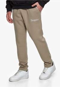 Beige sweatpants with elastic waist and side pockets, featuring "Dropsize" embroidered in white. Paired with white sneakers.