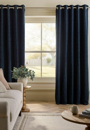 Großes Fenster, eingerahmt von dunkelblauen Vorhängen, mit einem beigen Sofa, einem runden Holztisch mit einer Blattpflanze und einem Couchtisch mit einer Tasse und Büchern.