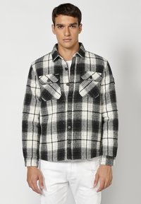 Zwart-wit geruite flanellen shirt met twee borstzakken, knoopsluiting en contrasterende textuur, gedragen over een wit T-shirt.