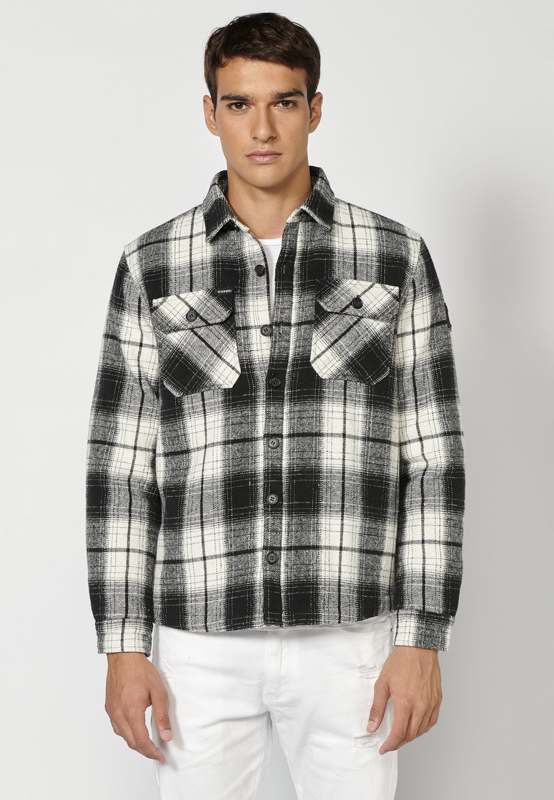 Zwart-wit geruite flanellen shirt met twee borstzakken, knoopsluiting en contrasterende textuur, gedragen over een wit T-shirt.
