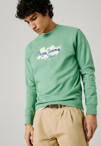 Mintgrüner Sweatshirt mit einem Logo in Blau und Weiß, Rundhalsausschnitt und gerippten Bündchen, kombiniert mit beigen Hosen.