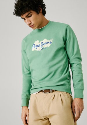 Mintgroene sweatshirt met een logo in blauw en wit, ronde halslijn en ribgebreide manchetten, gecombineerd met beige broek.