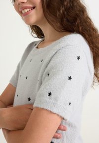 Lysegrå kortærmet sweater lavet af blødt, fuzzy stof, med sorte stjernemønstre over overfladen og fine ribbede manchetter.