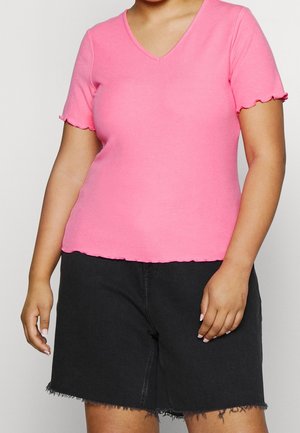 Kobieta nosząca jasnoróżowy krótki top z dekoltem w serek z falbankami oraz czarne, postrzępione krótkie spodenki jeansowe do kolan.