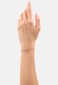 Braccialetto delicato con un piccolo ciondolo a forma di cuore adornato con cristalli, su una catena sottile, indossato su una mano sollevata su uno sfondo neutro.