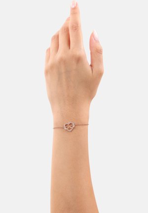 Braccialetto delicato con un piccolo ciondolo a forma di cuore adornato con cristalli, su una catena sottile, indossato su una mano sollevata su uno sfondo neutro.