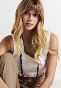 Jeune femme aux longs cheveux blonds portant un haut sans manches texturé blanc, un pantalon marron et un long collier de perles foncées, assise contre un fond uni.