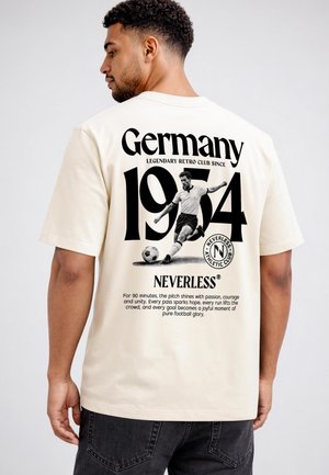Mann trägt ein cremefarbenes Hemd mit schwarzem Text und einer Grafik eines Fußballspielers, der "Germany 1974 Neverless Athletic Club" auf dem Rücken zeigt.