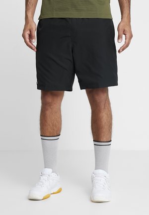 Homme portant un short de sport noir, des chaussettes grises avec des rayures noires, des baskets blanches, et un t-shirt vert olive, debout sur un sol gris.