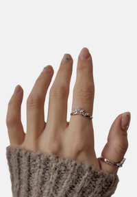 Srebrny pierścionek z kwiatowym wzorem, z centralnym diamentem i mniejszymi kamieniami, noszony na dłoni w teksturowanym, szarym swetrze.