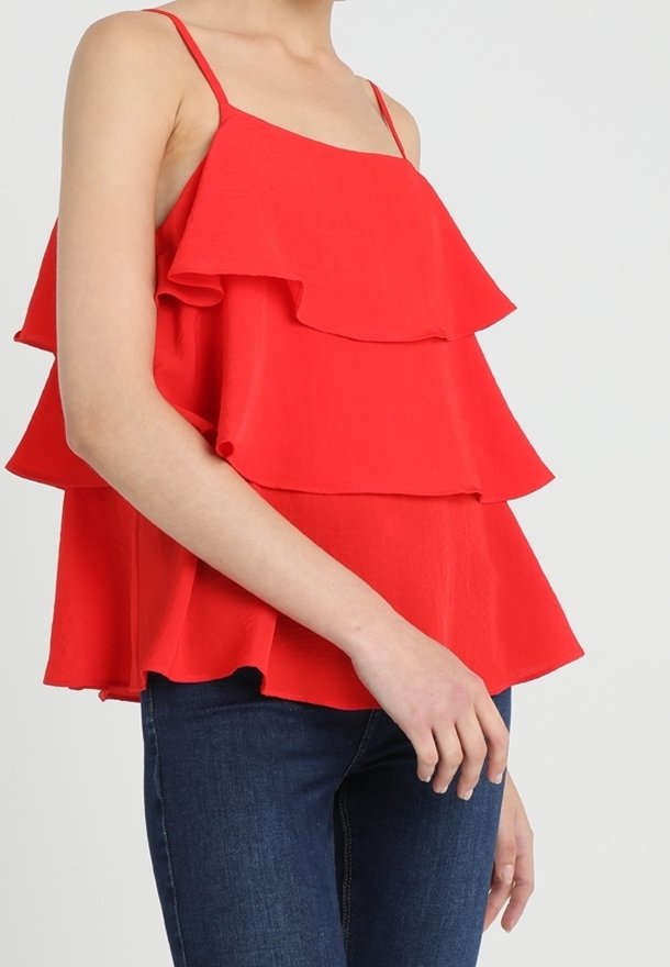 Vrouw in een felrode gelaagde mouwloze top met dunne bandjes en donkerblauwe jeans tegen een effen achtergrond.