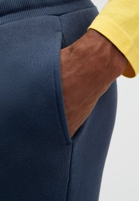 Hand in der Tasche einer marineblauen Jogginghose, mit sichtbarem gelbem Ärmelbündchen eines Langarmshirts vor einem schlichten hellen Hintergrund.
