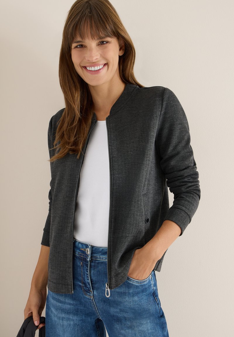 Giacca grigia a spina di pesce con zip frontale, design aderente e polsini a costine, abbinata a una maglietta bianca e jeans blu.