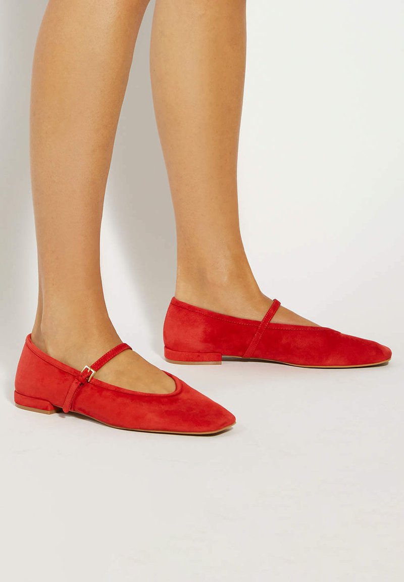 Voeten in rode suède Mary Jane ballet flats met kleine hakken en enkelbandjes op een lichte achtergrond.