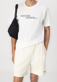 Weißes Baumwoll-T-Shirt mit grauem und schwarzem Logodruck, kombiniert mit cremefarbigen Shorts. Schwarze Umhängetasche mit Reißverschluss.