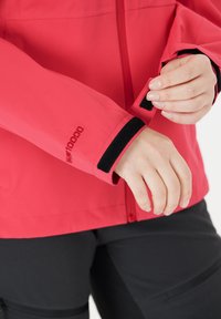 Red waterproof jacket with black Velcro wrist closure. Features a smooth texture and a front zipper. Hand adjusting cuff seen.