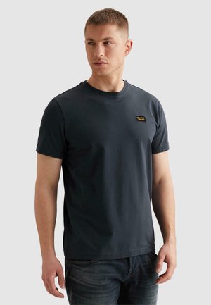 Homme portant un t-shirt à manches courtes bleu marine foncé avec un petit logo sur la poitrine et un jean bleu foncé, regardant légèrement sur le côté.