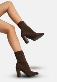 Braune gestrickte Knöchelstiefel mit einem gerippten Bund, spitzer Spitze und einem groben quadratischen Absatz. Glatte Textur mit kontrastierender matter schwarzer Sohle.