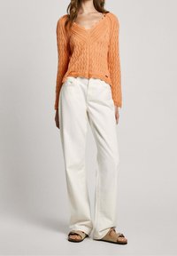 Oranje gebreide trui met een V-hals en geschulpte randen, gecombineerd met witte broek met wijde pijpen en bruine sandalen. Gebreide textuur met een patroon.