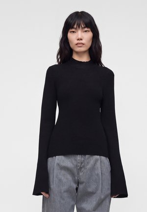 Donna con capelli lunghi e neri che indossa una maglia nera aderente a manica lunga con polsini a campana e pantaloni grigi larghi su uno sfondo semplice.