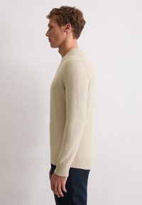 Maglione beige chiaro con una superficie texturizzata e polsini a costine, caratterizzato da un semplice collo rotondo e una silhouette aderente, indossato sopra pantaloni scuri.
