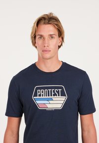 Camiseta de color azul marino hecha de algodón, con un diseño gráfico de olas y la palabra "PROTESTA" en blanco y colores pastel. Cuello redondo, mangas cortas.