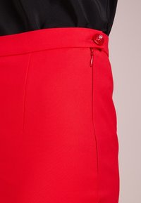 Červené přizpůsobené kalhoty s hladkým povrchem, vybavené bočním zipem a výrazným kulatým červeným knoflíkem.