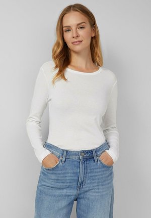 Balta garām piedurknēm krekls ar apaļu apkakli, apvienots ar gaiši ziliem augsto jostasvietu džinsiem. Mīksta, gluda auduma; ikdienišķs dizains.