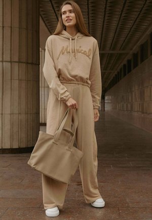 Fiatal nő bézs színű hétköznapi ruhában és fehér tornacipőben beltéren áll, kezében egy nagy bézs táska kézi fogantyúkkal.