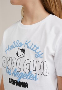 Camiseta de algodón blanca con un gráfico de "Hello Kitty" en negro y azul claro, mostrando el texto "Social Club Los Ángeles California". Tejido suave.