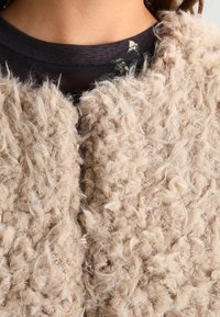 Veste en fausse fourrure bouclée beige portée par-dessus un top transparent à motif floral noir, ne laissant visible que le cou et la partie supérieure de la poitrine.